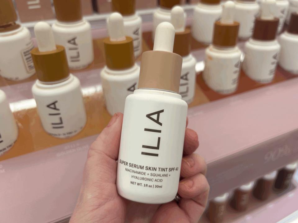 A hand holds a white bottle with Ilia logo and a tan dropper in Sephora. Other bottles are on display on a pink shelf in the background