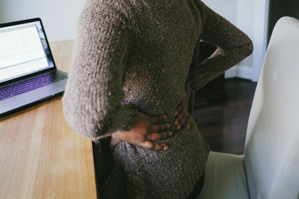 A person stands next to a desk with a laptop, holding their lower back in pain with both hands