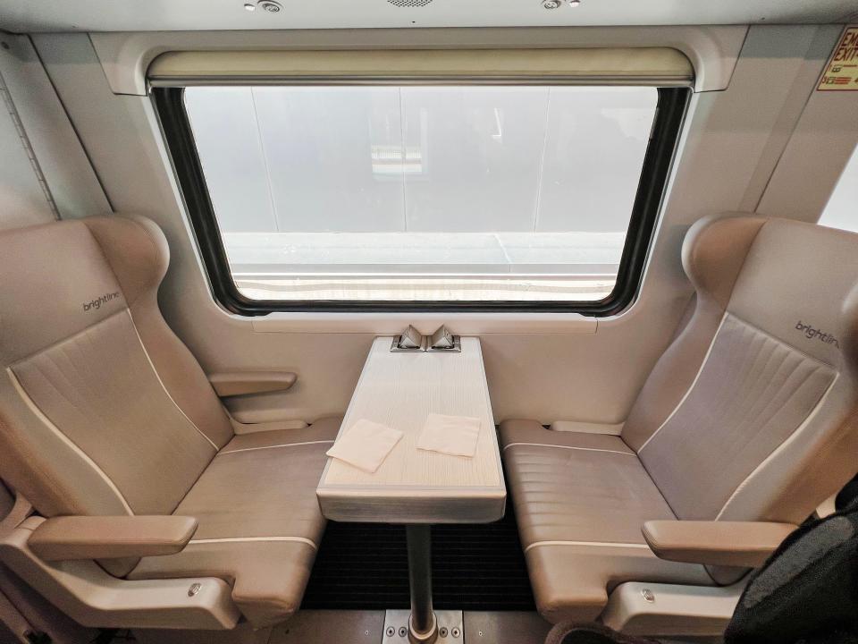 Tan, premium seats on the Brightline train are shown. Both are empty, facing one another with a small table between them.
