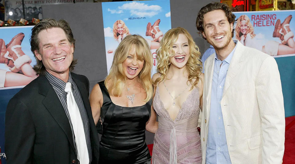 Kurt Russell in a black suit and black and white striped shirt laughs with Goldie Hawn ina. black dress, Kate Hudson in a light pink lacy dress and Oliver Hudson in a cream jacket on the carpet