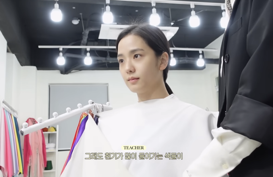 Jisoo is being assisted by a person in a suit with a clothes hanger in a well-lit changing room. Korean text is at the bottom of the image