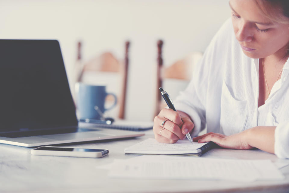 Woman writing on a pad working at home sitting at the dining room table. There is a laptop computer and mobile phone on the table.