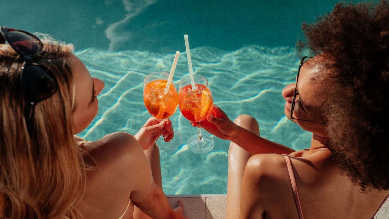 two female friends seen from above and behind poolside clinking plastic glasses with straws in them