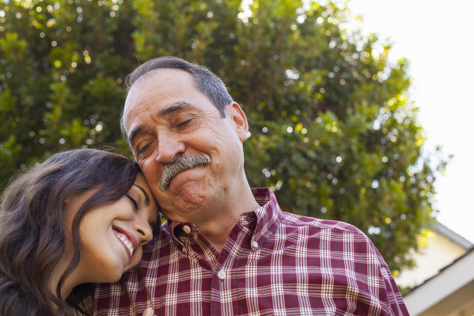 A young woman leans her head on an older man's shoulder. Both are smiling warmly. They are outdoors with trees in the background