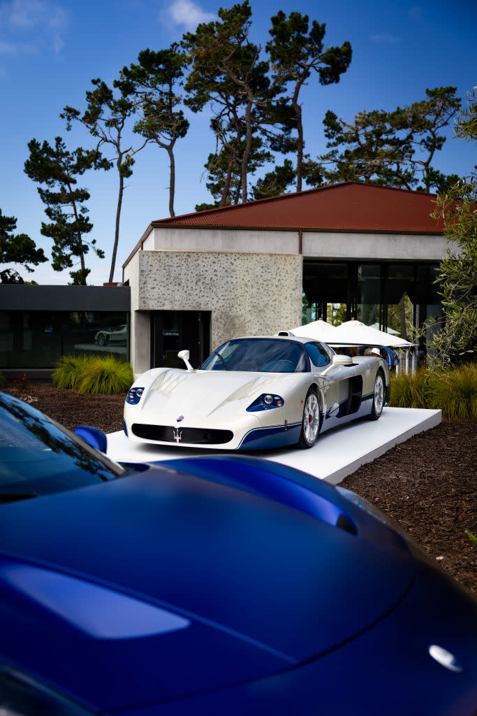 A Maserati sports car is displayed outdoors on a white platform with trees and a modern building in the background