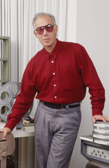 A man posing in gray sunglasses, gray pants and a red, button-up shirt