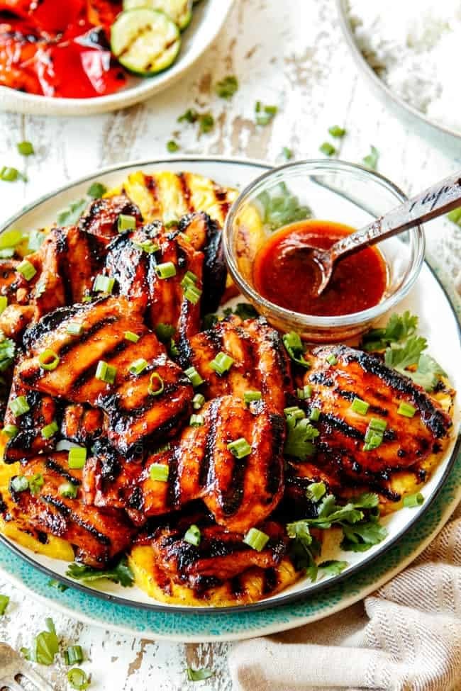 Grilled chicken thighs garnished with chopped green onions, served with a bowl of barbecue sauce on a plate with a side of grilled vegetables