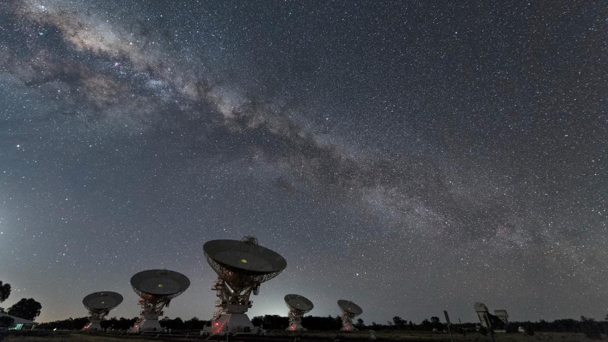  satellite dishes at the bottom of the image are pointed toward a star-studded sky with the bulge of the Milky Way stretching across the center of the image.  