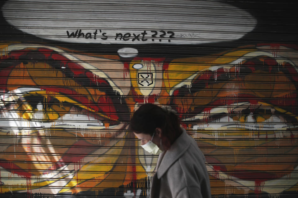 A woman wearing a face mask to protect against coronavirus, passes in front of graffiti painted on the shutter of a closed restaurant during a lockdown order by the Greek government to control the spread of the virus, in Athens, Monday, March 30, 2020. Greece's prime minister is calling on all his cabinet ministers and the lawmakers of his center-right New Democracy party to donate 50% of their salaries over the next two months to the fight against the spread of the new coronavirus. (AP Photo/Thanassis Stavrakis)