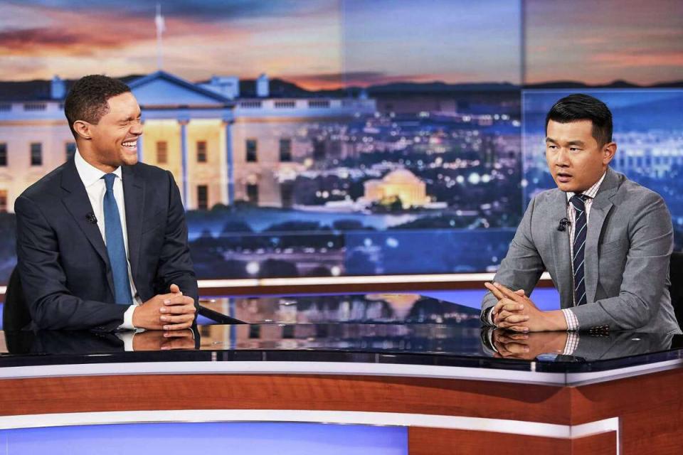 Trevor Noah laughing while Ronny Chieng is speaking at a desk