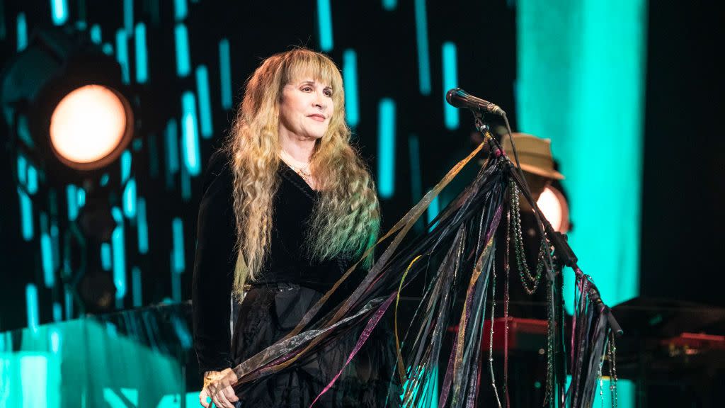 stevie nicks stands onstage behind a microphone decorated with strips of fabric and beads, she looks out at an unseen crowd with a soft smile and wears a black outfit