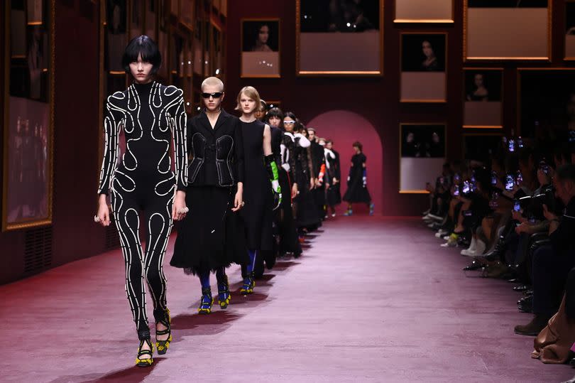 Models dressed in all black couture fashion walking in single file on a catwalk