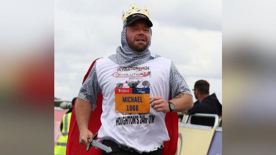 Michael running wearing a knight's costume with a red cape and carrying a plastic sword. His name is on his T-shirt