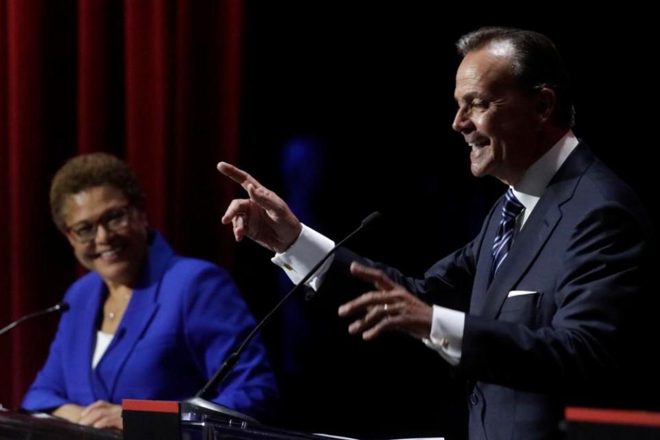 A man and a woman stand at individual podiums. The woman is smiling and looking at the man who is speaking and pointing with his right hand.