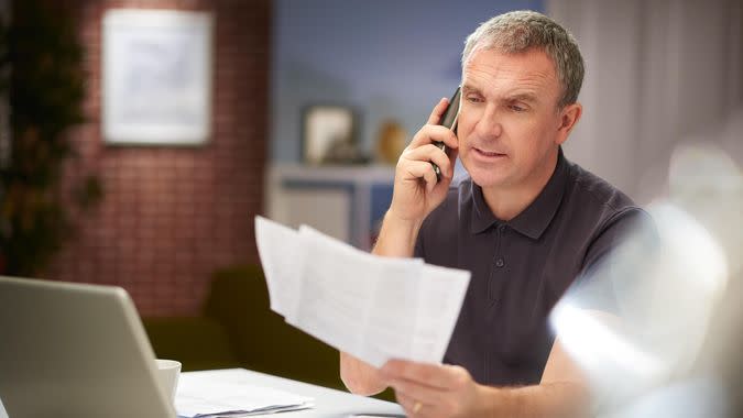 a mature man sits at his dining table at home looking through some bills and paperwork , whilst chatting on the phone .