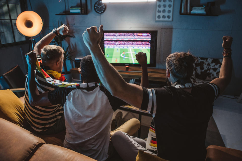 Three people with raised arms cheer while watching a soccer match on TV in a living room