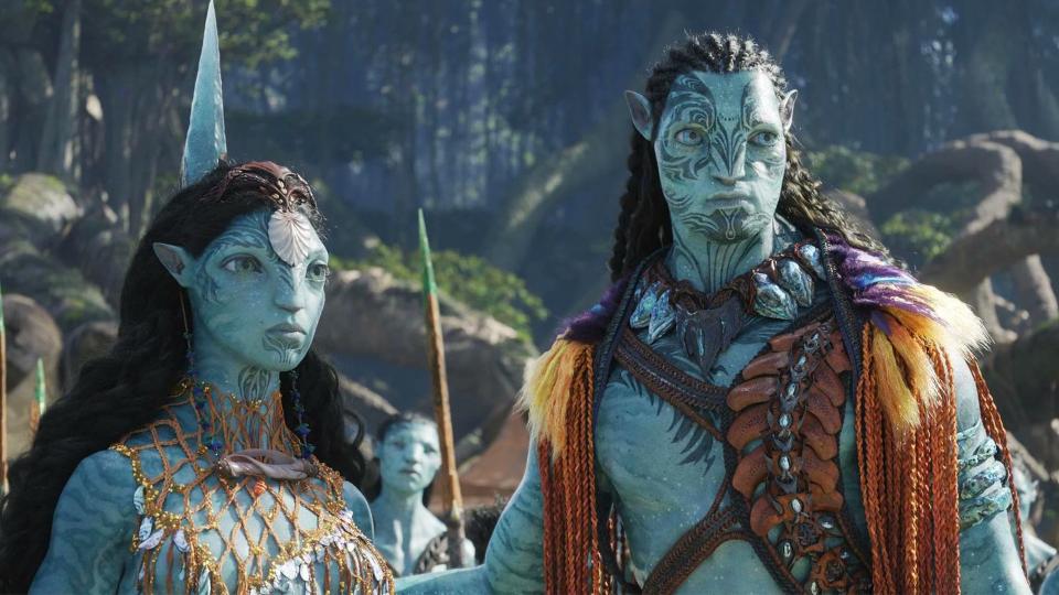 Still from the movie Avatar: The Way of Water (2022). Here we see two tall blue pi-pedal creatures called Na'vi (female on the left, male on the right). They both have blue skin with dark blue stripes and long black hair. They have triable tattoos on their face and are wearing some ocean-themed garments.