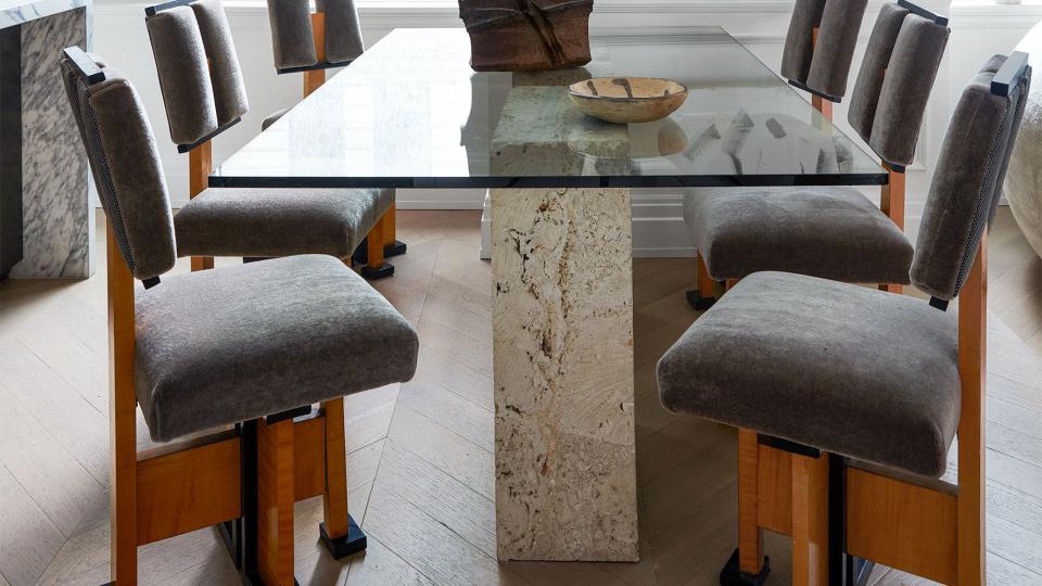 a room with a marble pedestal base table and gray suede chairs with wood frames and a black and white abstract painting on the far wall next to window