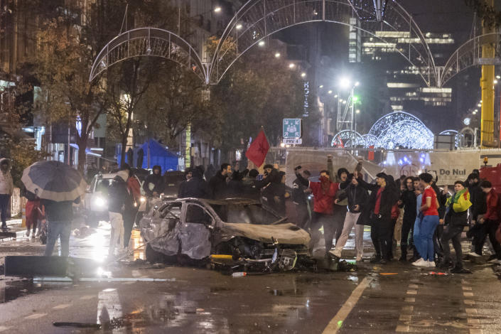 Moroccan soccer supporters cheer as they stand next to a burned out car in Brussels, Sunday, Nov. 27, 2022. Police had to seal off parts of the center of Brussels and moved in with water cannons and tear gas to disperse crowds following violence during and after Morocco's 2-0 win over Belgium at the World Cup. (AP Photo/Geert Vanden Wijngaert)