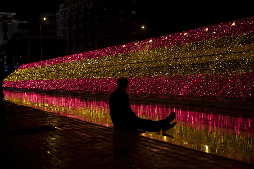 A man sits in front of a bank of Christmas lights in the colors of the Spanish flag in Madrid, Spain, Friday, Nov. 27, 2020. People are waiting to see what restrictions will be in place over Christmas and the New Year as Spanish regions are continuing to adjust their various limitations against the coronavirus pandemic. (AP Photo/Paul White)