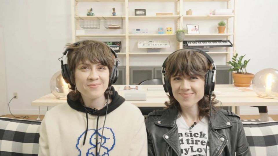 Two women smile at the camera as they wear headphones in front of a shelf background.