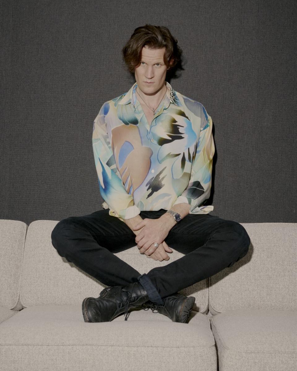 A man in black jeans and a colorful patterned shirt sits on the back of a sofa.