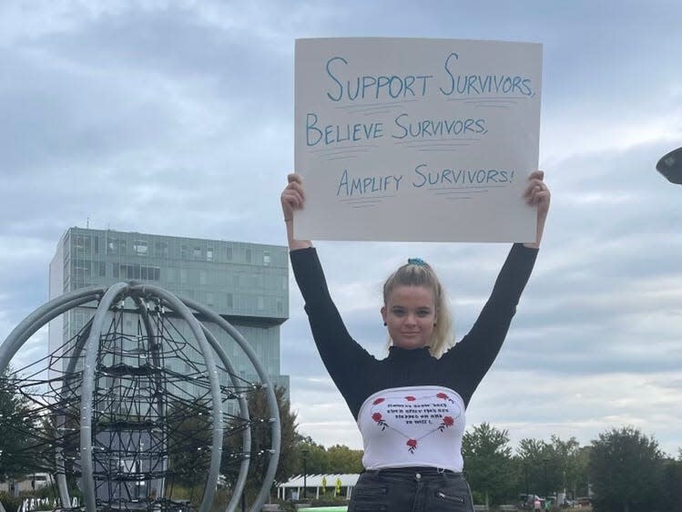 A girl in a long-sleeved white T-shirt with black sleeves holding up a sign that says, "Support survivors/ believe survivors / amplify survivors"