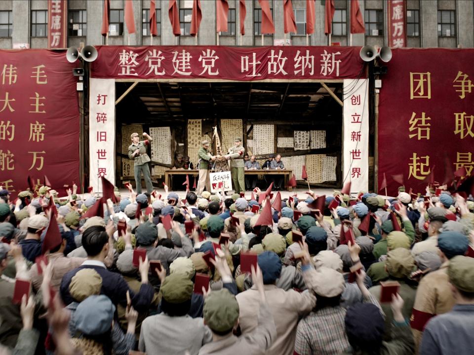 a struggle session during the chinese cultural revolution in three body problem, with text in mandarin heralding the beginning of a new age. hundreds hold up little red books as two red guards force a man down on stage