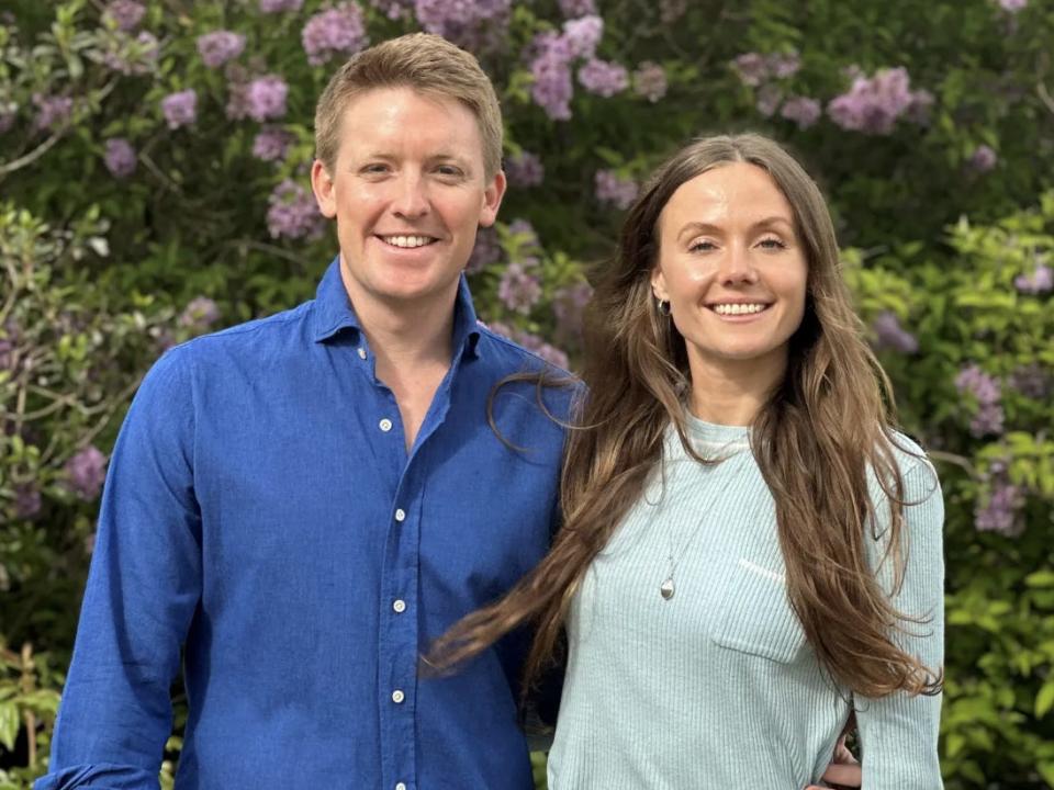 Hugh Grosvenor standing with his arm around Olivia Henson in front of a bush with pink flowers.