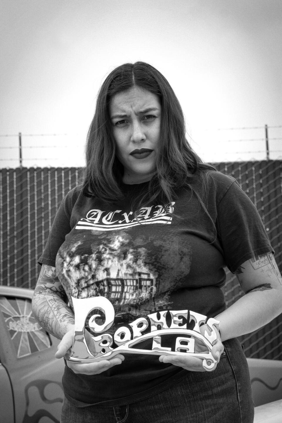 a black and white photo of Jacqueline Valenzuela holding up a "Prophets LA" lowrider plaque