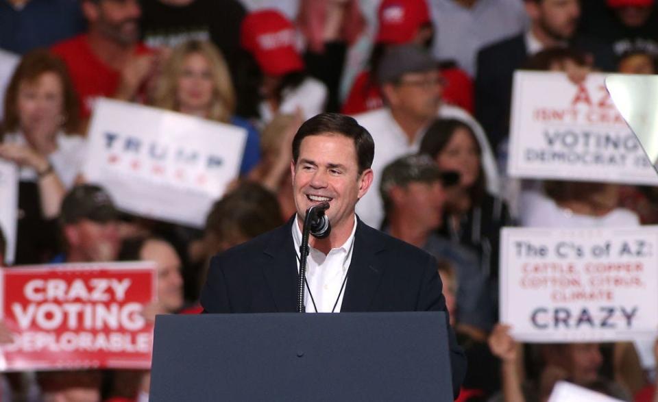 Arizona Governor Doug Ducey wears a black suit jacket and speaks into a microphone on a stage.