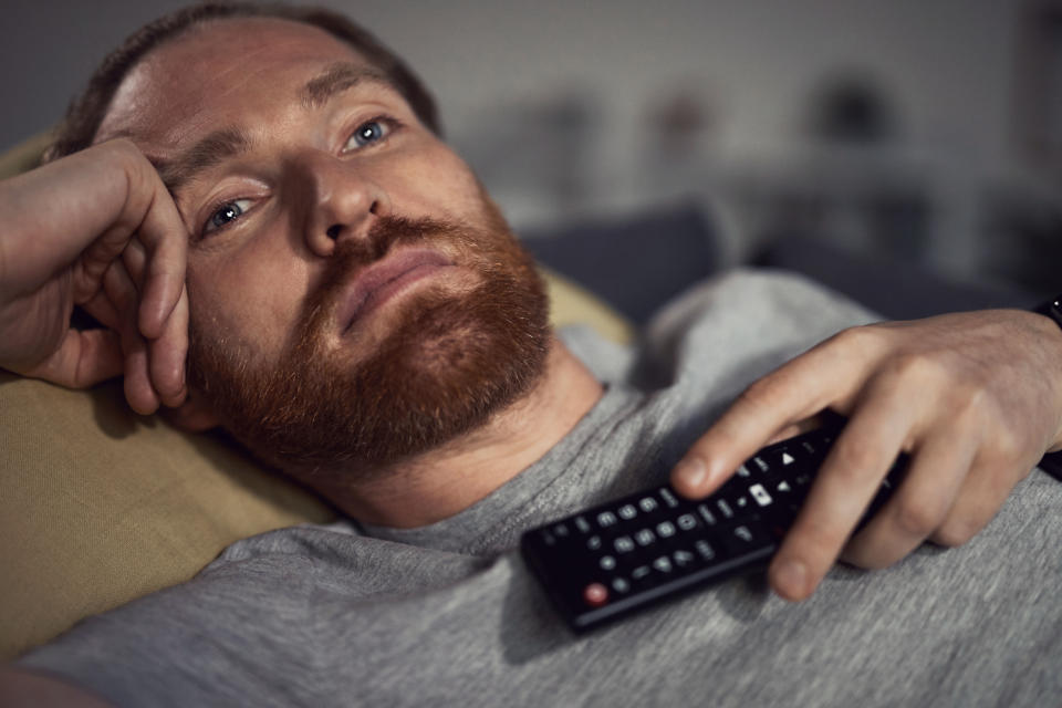 Portrait of bearded adult man watching TV at night while lying on couh in dark room and switching channels, copy space