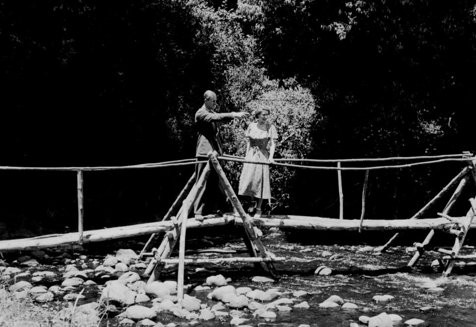 A man in a suit points as a woman on the right looks on. Both are standing on a bridge