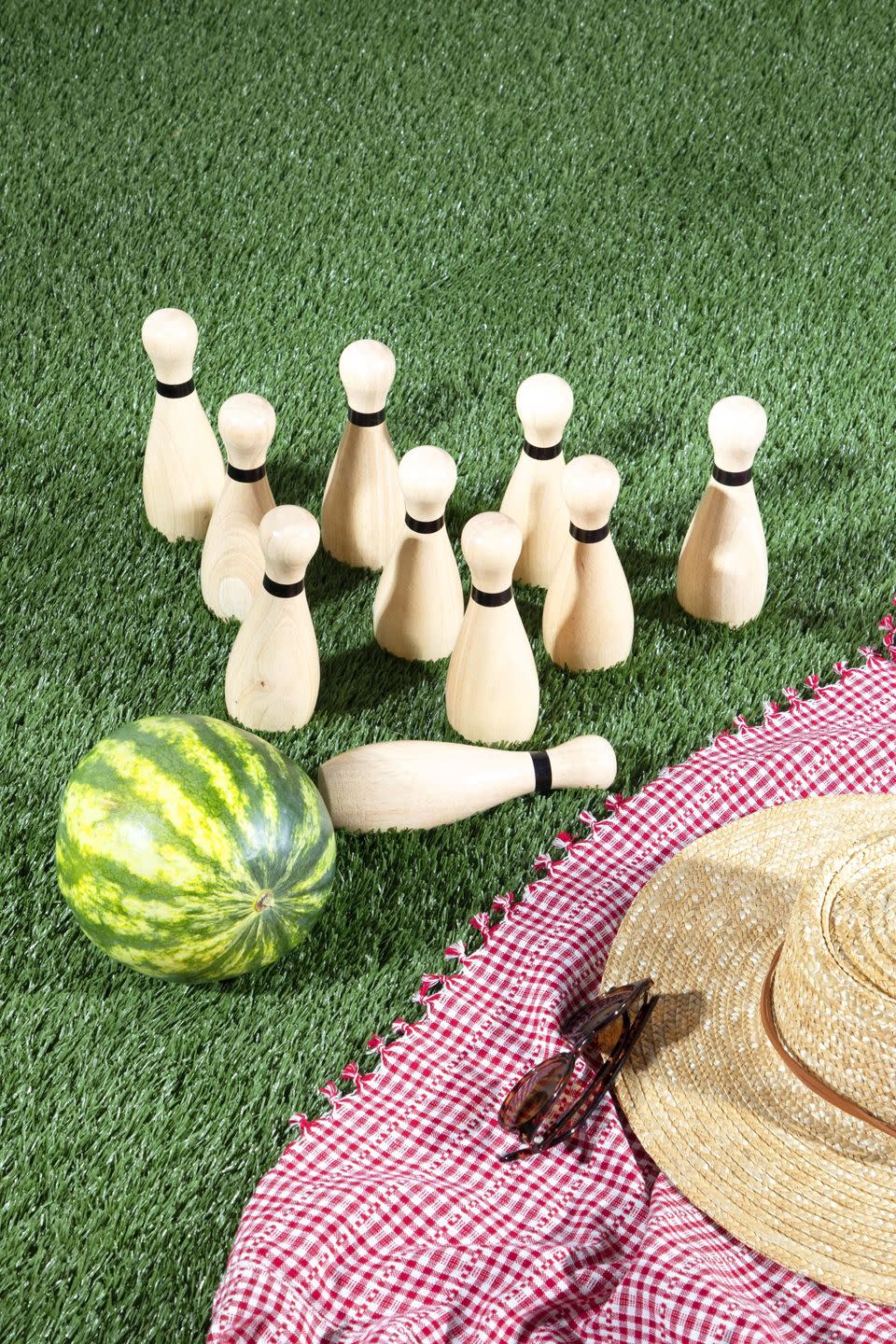 watermelon and bowling pins on the lawn next to a red and white gingham picnic blanket, straw hat, and sunglasses