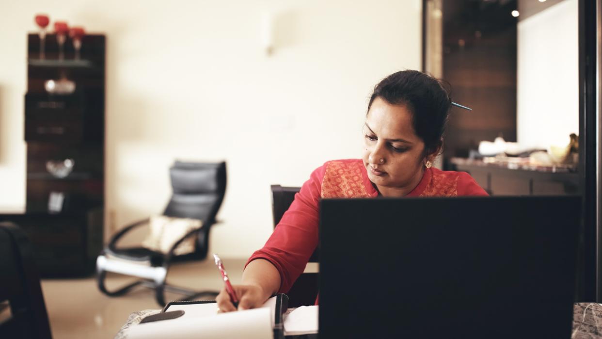 Mid-age woman working at home, using laptop and writing in notepad.