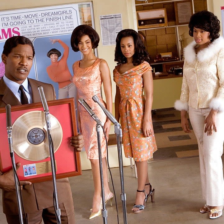 man in front of microphones holding a record while three women stand behind him