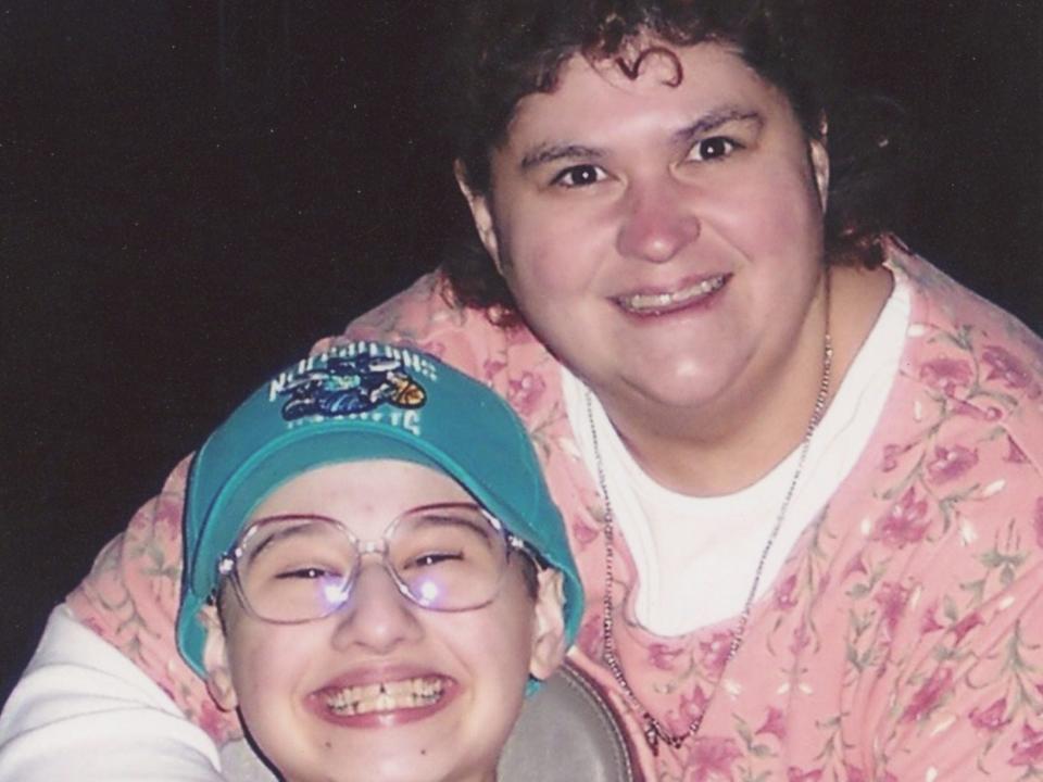 dee dee blanchard holding her arms around gypsy rose blanchard, who is smiling widely, sitting in a chair, and wearing a hat on her head