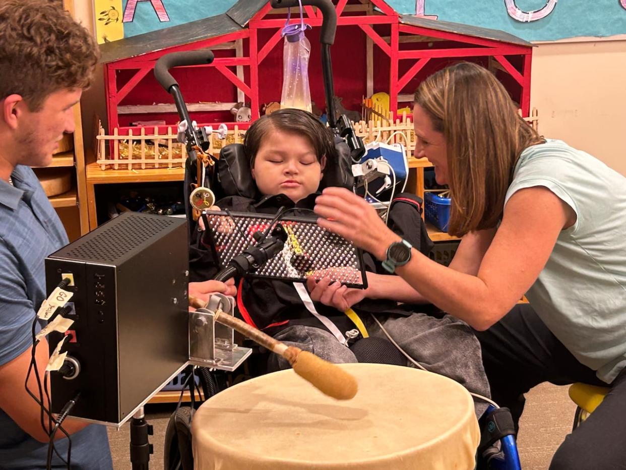 Gerry Lewis plays his custom-made drum Tuesday with the help of his occupational therapist Jennifer Neill and UPEI engineering student Christian d'Entremont. (Laura Meader/CBC - image credit)