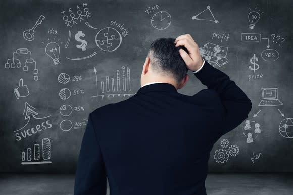 A person scratches his head while looking at diagrams on a chalkboard.