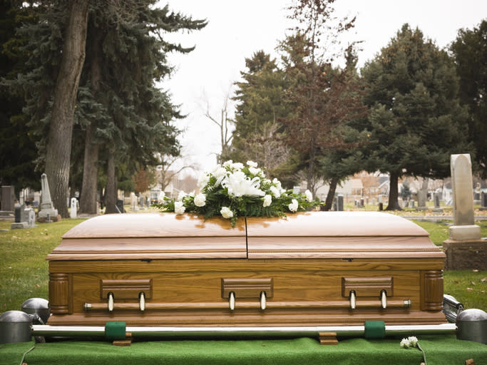 A closed coffin adorned with white flowers set up for a burial in a cemetery, surrounded by tombstones and trees