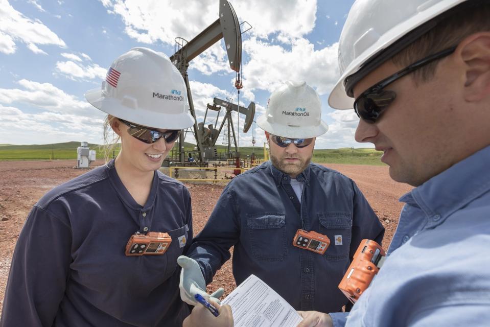 Three people wearing Marathon Oil hard hats looking at documents at an oil well site.