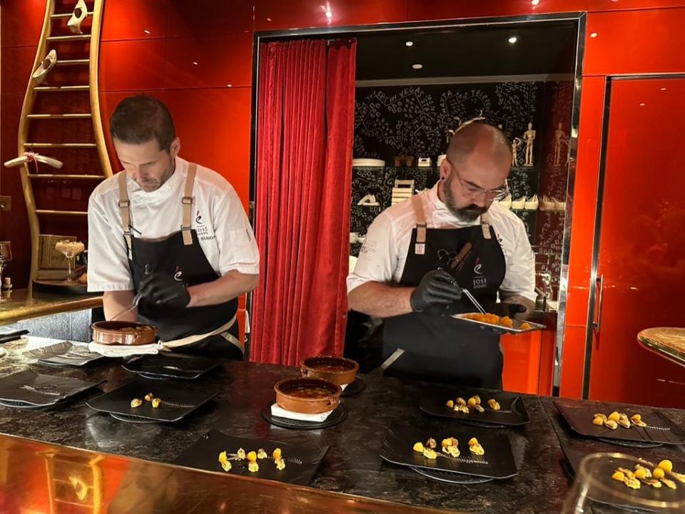 Two chefs with gloves on meticulously constructing plates in front of a red backdrop