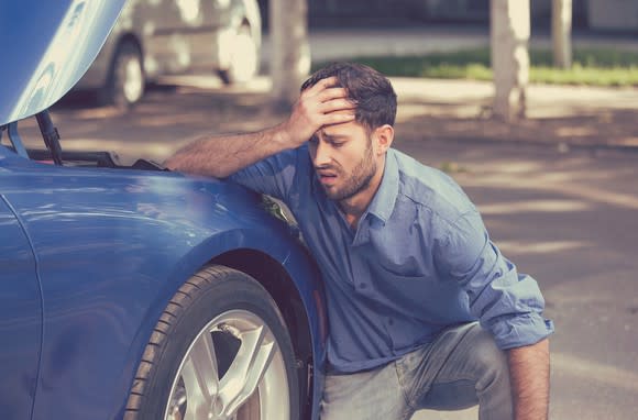 Man holding his head in exasperation while kneeling next to a car with its hood raised.