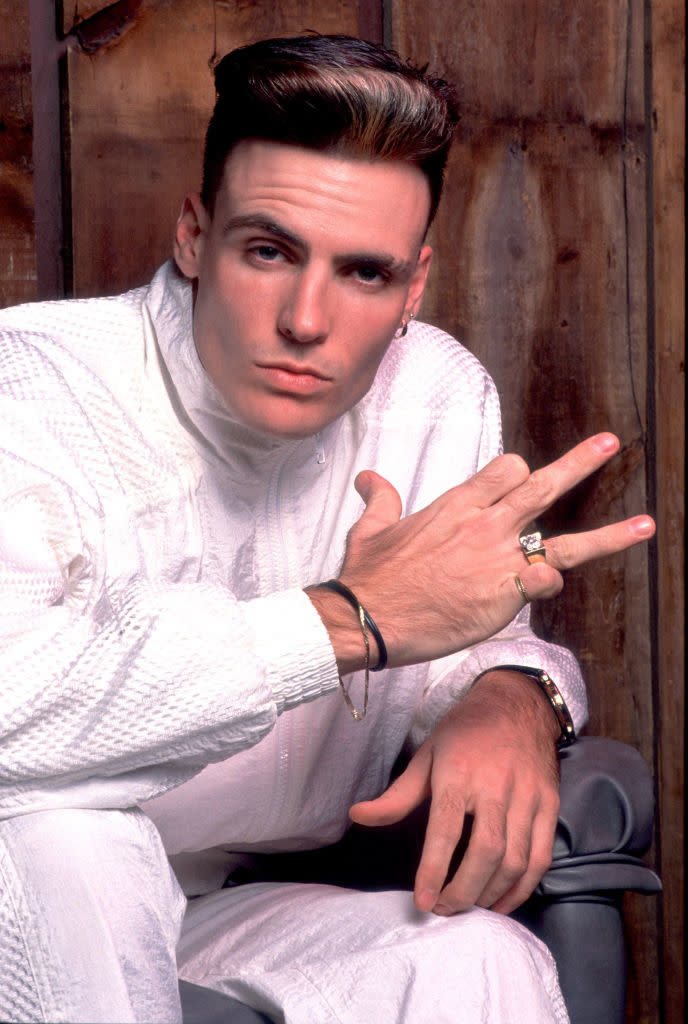 Vanilla Ice in a white textured outfit, striking a hand gesture with two fingers extended and one bent