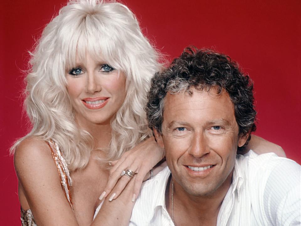 Actress Suzanne Somers and husband Alan Hamel pose for a portrait in front of a red background wearing 80's glam clothing.