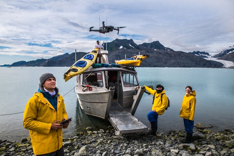 Researchers in yellow jackets pilot a drone next to a moored research vessel.