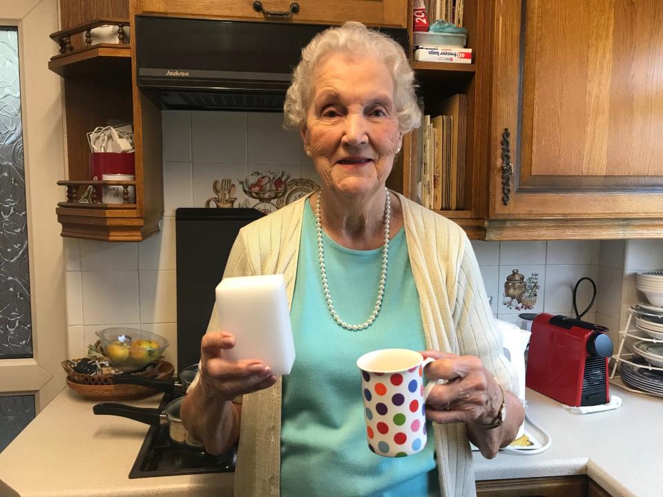 Image of an older woman with short curly white hair holding a mug with a colorful polka dot design in one hand and a Magic Eraser in the other. She wears a turquoise t-shirt, a white ribbed cardigan, pearl earrings, and a pearl necklace. Behind her is a kitchen stove, with wooden cabinets on the white tiled wall.