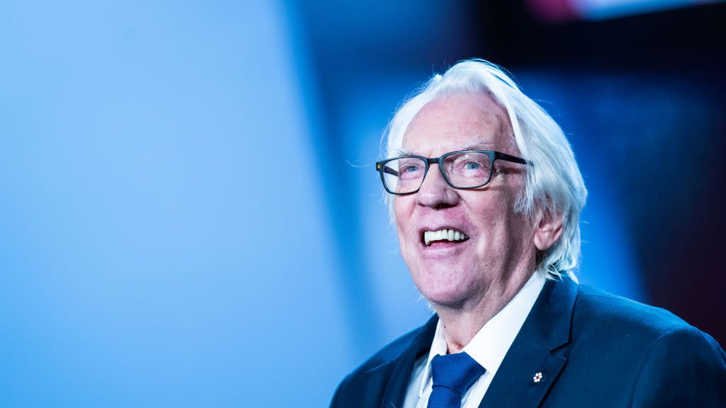 donald sutherland smiles and wears black framed glasses, a navy suit jacket, royal blue tie and white dress shirt