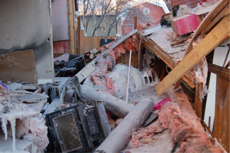 A photo from the fire investigation report showing the right side of the home on Dagenais Drive in Yellowknife after a propane explosion in December 2022. A wood stove is seen in the bottom left corner and beyond it — a toppled propane tank from Matonabee Petroleum. (N.W.T. Office of the Fire Marshal - image credit)