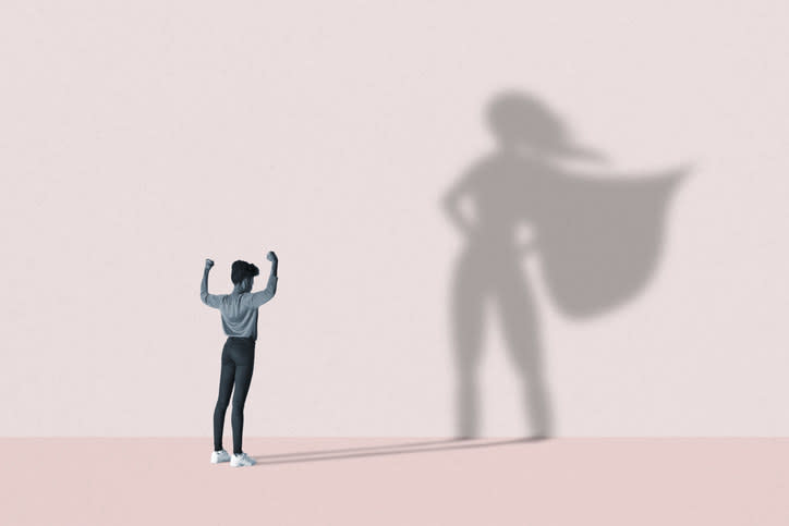 Young woman flexing muscles in front of large superhero shadow on pink background